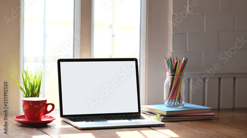 Photo of white blank screen computer laptop putting on working desk with bunch of flowers, notebook and pencil holder over comfortable living room as background. Orderly workspace concept. photo
