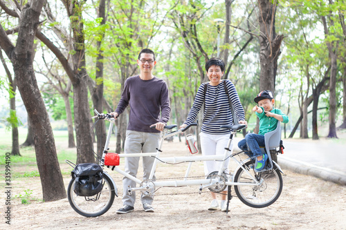 Portrait of good looking & happy Asian family (father, mother and son) with white tandem bicycle in beautiful city urban park. Dad and mom stand and smile, little boy sit in child seat on the back.