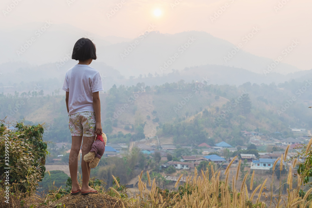 Fototapeta premium little girl holding a teddy bear looking her future on mountain at sunset.
