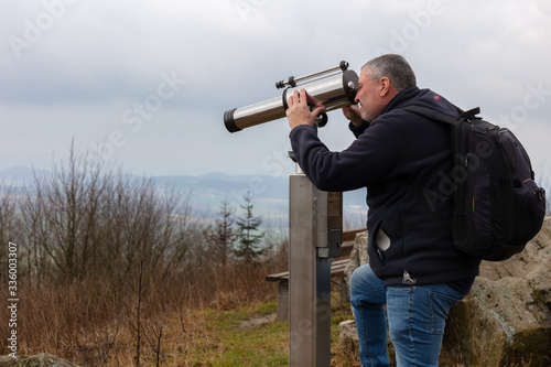 A hiker with a backpack stands on the highest mountain in eastern Westphalia in Germany and looks into the distance through binoculars. The place is called Köterberg. It is a cloudy winter day.