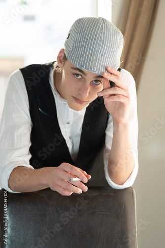 A young man in a cap and vest smokes by the window