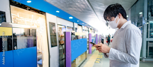 Asian man wearing surgical face mask using smartphone at skytrain station platform. Wuhan coronavirus (COVID-19) outbreak prevention in public transportation. Health awareness for pandemic protection