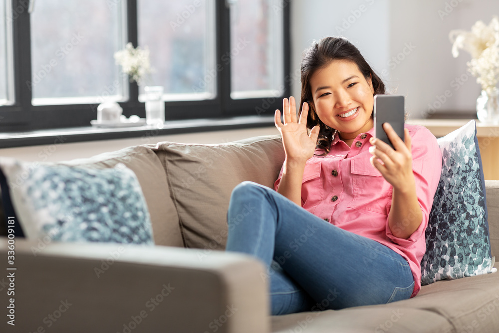 people, technology and communication concept - happy smiling asian young woman in pink shirt sitting on sofa and having video call on smartphone at home