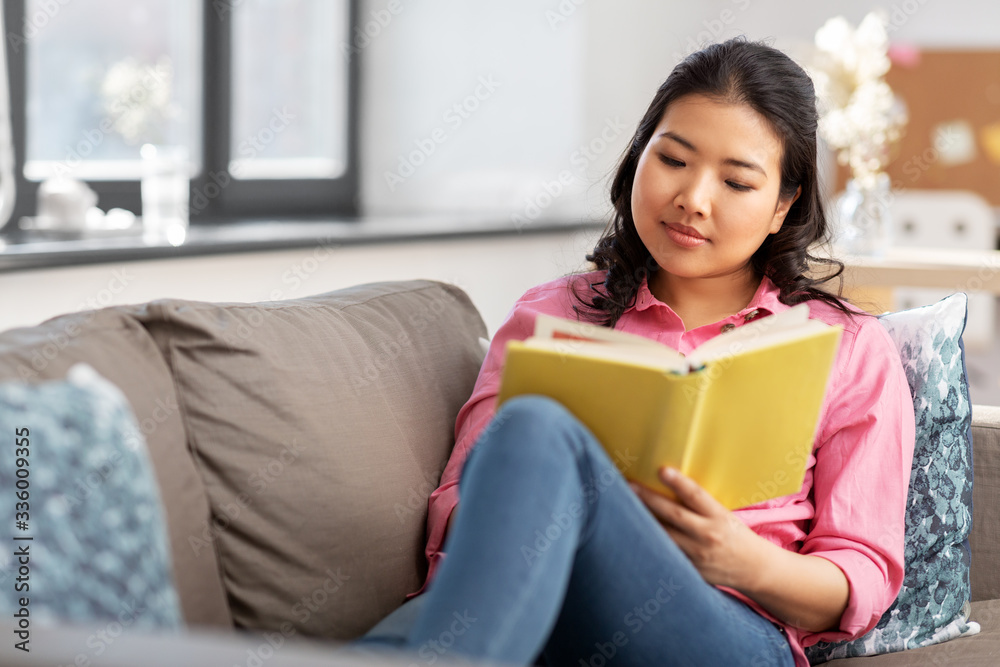 people and leisure concept - happy asian young woman in pink shirt sitting on sofa and reading book at home people and leisure concept - happy asian young woman in pink shirt sitting on sofa and reading book at home