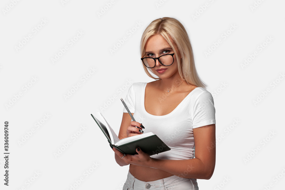 Young blonde woman wearing casual t-shirt standing over isolated white background with a notebook notepad. Portrait of girl with perfect face and skin with glasses. Mockup. Young blonde woman wearing casual t-shirt standing over isolated white background with a notebook notepad. Portrait of girl with perfect face and skin with glasses. Mockup.