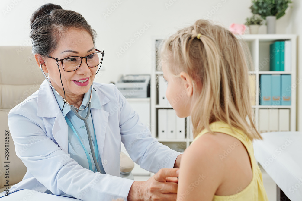 Smiling mature Asian female pediatrician checking heartbeat of little patient Smiling mature Asian female pediatrician checking heartbeat of little patient
