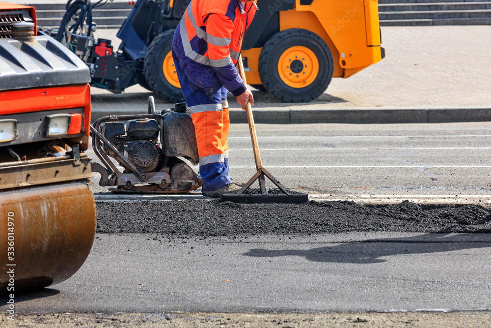 A road worker is leveling fresh asphalt on a road section against a background of road equipment.