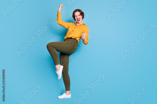 Full length photo of pretty business lady raise fists celebrating startup successful investment income wear yellow shirt green pants footwear isolated bright blue color background