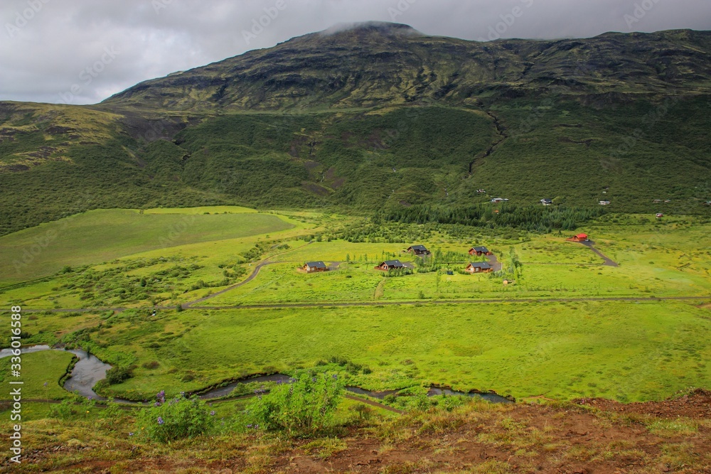 small houses are located in a valley at the foot of green hills, near the winding river flows, the sky is overcast, the nature of Iceland, a summer day