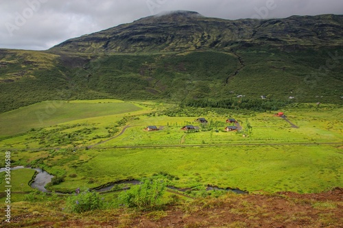 Fototapeta Naklejka Na Ścianę i Meble -  small houses are located in a valley at the foot of green hills, near the winding river flows, the sky is overcast, the nature of Iceland, a summer day