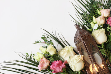 Vintage bronze Moroccan, Arabic lantern with glowing candle, green branches, pink rose flowers and palm leaves on white table background. Greeting card for Muslim holiday Ramadan Kareem.