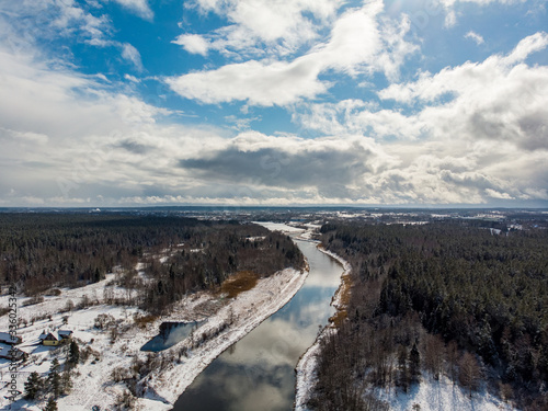 Drone areal view of river Venta flowing near forest on a cold snowy winter day.