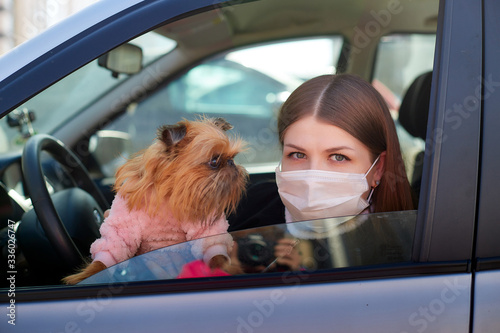 Girl in white medical gauze mask in the car with a dog in sunny spring day. Protection against disease and coronavirus during epidemics and pandemics. Covid-19 in Russia and russian city