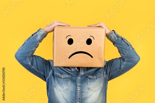 Hombre con caja de cartón en la cabeza con gesto pensativo preocupante y manos sosteniendo la cabeza sobre fondo amarillo liso brillante y aislado. Vista de frente y de cerca. Copy space