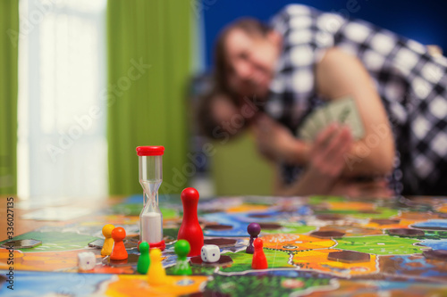 Board game concept- friends spend time together. Board game field, figures, dice, coins and sandglass. Two people play holding cards on blurred background