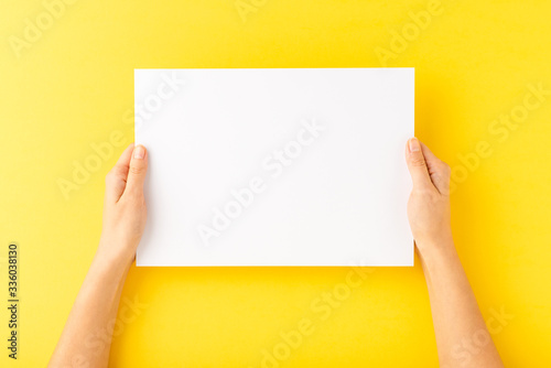 Woman’s hands holding blank a4 paper sheet on yellow table. Close up