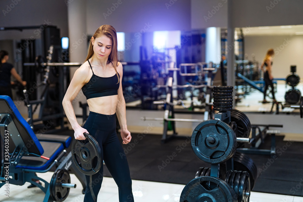 Fototapeta premium Athletic woman holds a black pancake rod and looks at camera in the gym in the sport equipment background. Health life concept.