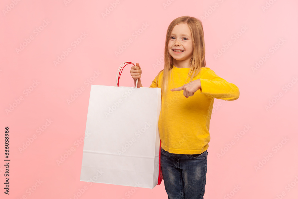 Look at my purchase! Portrait of charming happy little girl pointing at shopping bags and smiling at camera, satisfied with Black Friday discounts, low prices. studio shot isolated on pink background Look at my purchase! Portrait of charming happy little girl pointing at shopping bags and smiling at camera, satisfied with Black Friday discounts, low prices. studio shot isolated on pink background