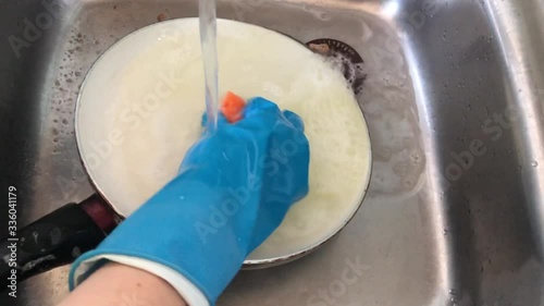 A woman in rubber gloves washes a dirty pan with a foam sponge in the sink. Washing dishes at home in the kitchen.