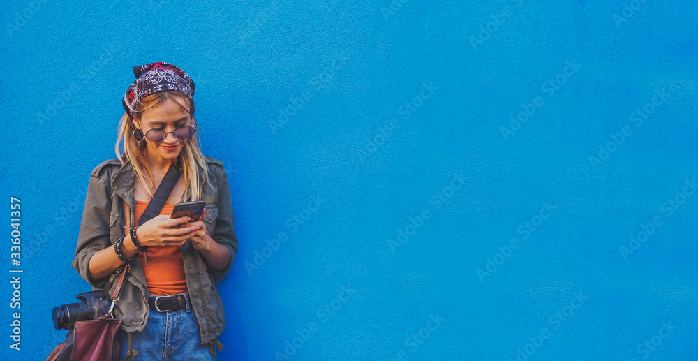 Young girl standing by the blue background holding a smartphone - A hipster stylish girly smiling and checking out social media