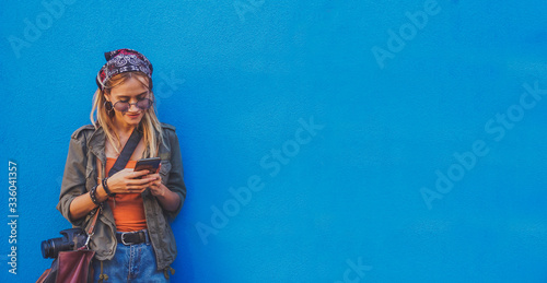 Young girl standing by the blue background holding a smartphone - A hipster stylish girly smiling and checking out social media