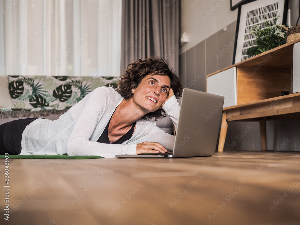 happy and smiling women lying on the floor on her stomach in her living room at home with a laptop typing and looking at in the camera happy and smiling women lying on the floor on her stomach in her living room at home with a laptop typing and looking at in the camera