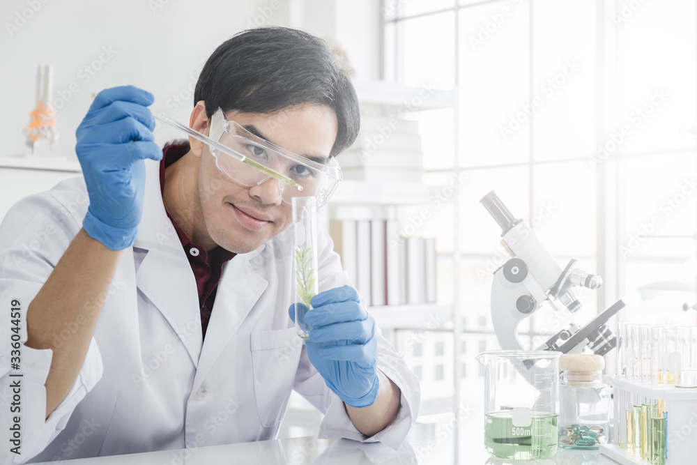 A male scientist with black hair wearing white coat and protective glassware dripping solution into a test tube with a green plant sample in a white laboratory or hospital. A male scientist with black hair wearing white coat and protective glassware dripping solution into a test tube with a green plant sample in a white laboratory or hospital.
