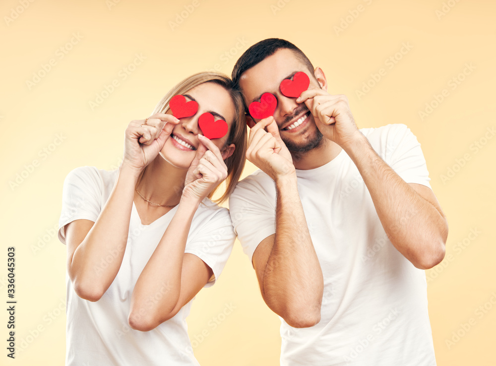 Happy couple have fun holding small paper hearts by their eyes over studio background. Happy couple have fun holding small paper hearts by their eyes over studio background.