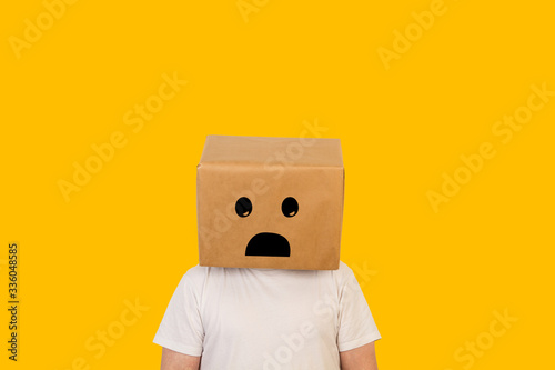 Hombre con caja de cartón en la cabeza con un gesto de tristeza en la cara sobre fondo amarillo liso brillante. Vista de frente. Copy space