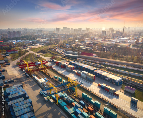 Aerial view of container loading and unloading at sunset. Top view of containers at logistics terminal in city, factory, train, railroad, sky. Business. Freight transportation containers on railway