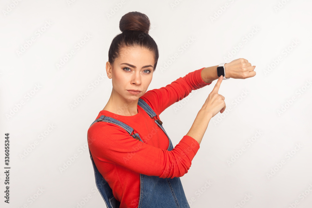 Too late, hurry up! Portrait of displeased impatient girl with hair bun in denim overalls pointing at wrist watch with warning expression, time to go. indoor studio shot isolated on white background Too late, hurry up! Portrait of displeased impatient girl with hair bun in denim overalls pointing at wrist watch with warning expression, time to go. indoor studio shot isolated on white background