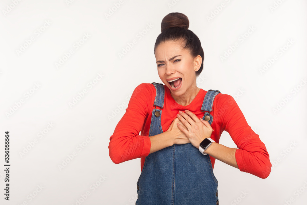 Cardiac problems. Portrait of young fatigued woman with hair bun in denim overalls clasping chest, feeling acute pain, heart attack from anxiety, infarction. studio shot isolated on white background Cardiac problems. Portrait of young fatigued woman with hair bun in denim overalls clasping chest, feeling acute pain, heart attack from anxiety, infarction. studio shot isolated on white background