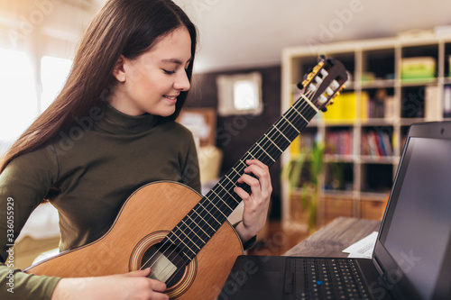 Focused girl playing acoustic guitar and watching online course on laptop while practicing at home. Online training, online classes.