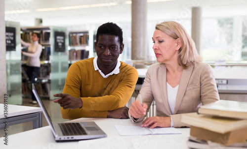 Friendly female tutor helping to diligent african-american man preparing for exam in library