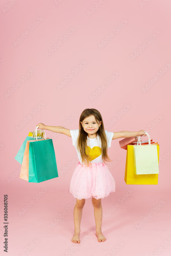 Little blonde girl enjoys her shopping on a pastel pink background with copyspace. Sale. Cute little girl with many multicolored shopping bags in studio