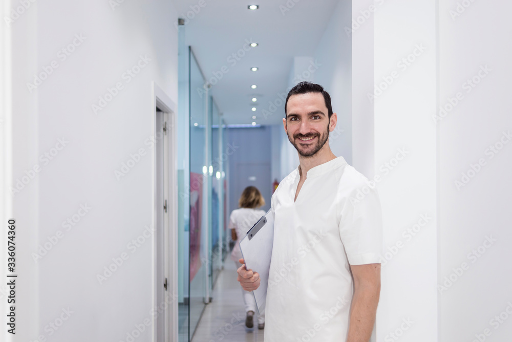 Portrait of a Young smiling bearded doctor holding a folder while leaning on white wall and looking to camera at a clinic corridor Portrait of a Young smiling bearded doctor holding a folder while leaning on white wall and looking to camera at a clinic corridor