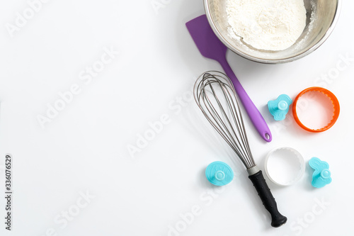 Kitchen items, spatula, whisk, baking dish, bowl of flour on a white background