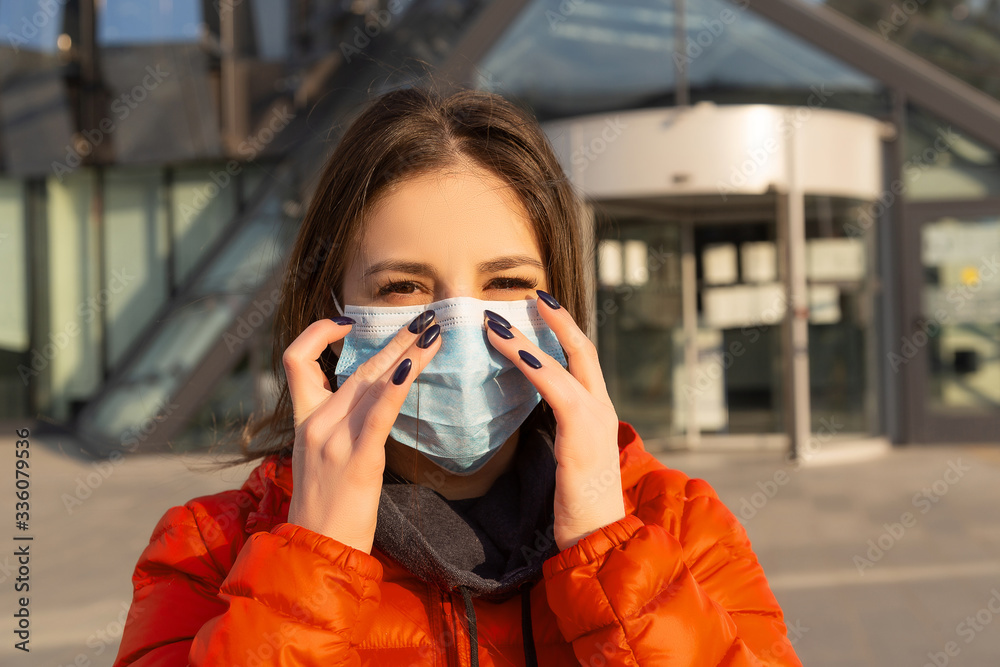 A woman wears a protective mask against allergies, viruses, air pollution. Climate change concept. Portrait of a young woman in a medical mask from a coronavirus in a red jacket on the street.