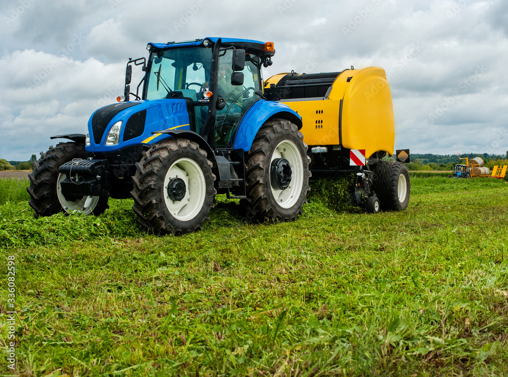 new blue tractor with baler in motion on farmland with cloudy sky new blue tractor with baler in motion on farmland with cloudy sky
