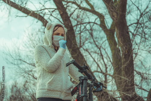 Young woman in medical mask and gloves coughing, holding on to rudder of bicycle in countryside. Female protecting yourself from diseases on walk. Concept of threat of coronavirus epidemic infection.