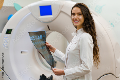 Medical student looks in camera and smile. Medical stands with x-ray picture of patient brain in her hands. woman stays in front of kt komputer tomograhphy. Girl in medical clinic gotes new expirience photo