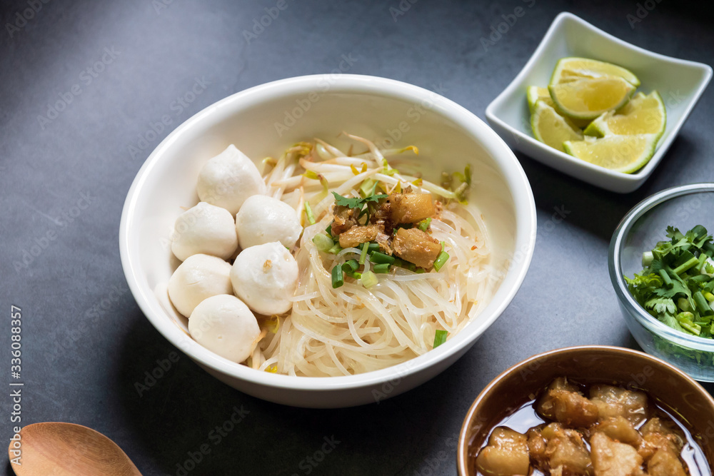 Rice stick noodles with fish ball in white bowl. Garlic cracklings with lard, lime and coriander with spring onion various ingredients and condiment on table. Rice stick noodles with fish ball in white bowl. Garlic cracklings with lard, lime and coriander with spring onion various ingredients and condiment on table.