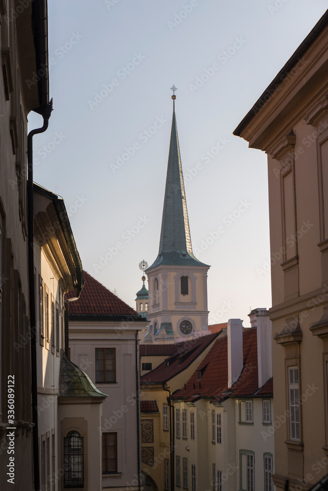 view of a Prague street, narrow street, light colored buildings with reddish roofs in the background highlights the pointed roof of a church