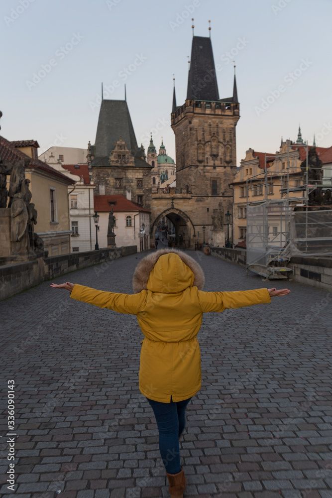 woman in yellow coat and open arms walking on the Charles Bridge in Prague at dawn, the bridge is empty and in the background we can see the typical buildings of the city with the pointed roofs