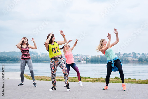 A group of young women, wearing colorful sports outfits, doing zumba exercises outside by city lake. Dancing training to loose weight in summer. Healthy lifestyle concept. Female sport leisure.