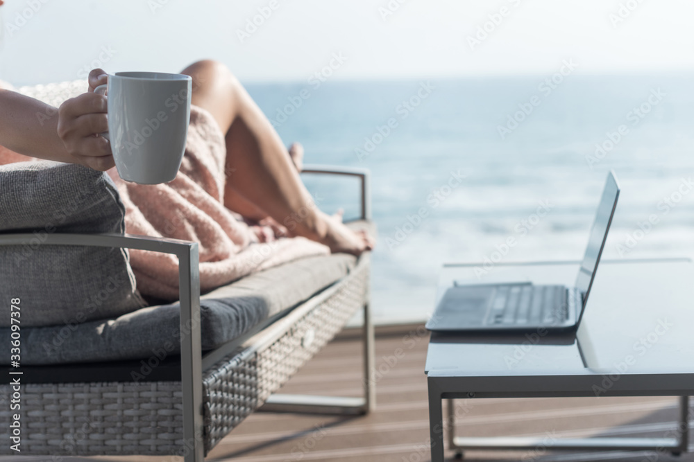 Obraz premium Beautiful brunette woman freelancer wrapped in a plaid resting on a couch and sunbathing on the terrace with a cup of coffee at summer day. Sea view. WFH. Working from home
