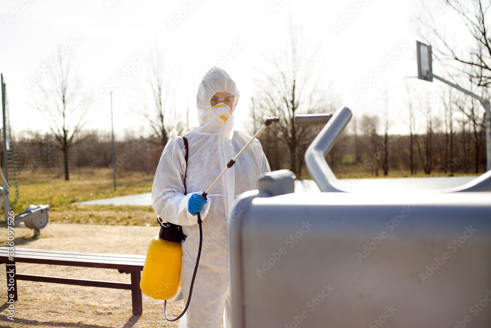 Naklejka premium Osoba w kombinezonie ochronnym spryskuje sprzęt na siłowni zewnętrznej by go zdezynfekować