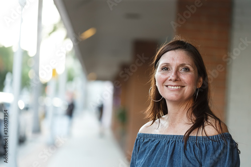 Happy and smiling middle aged 50+ retired woman having confidently standing alone on sidewalk in urban area