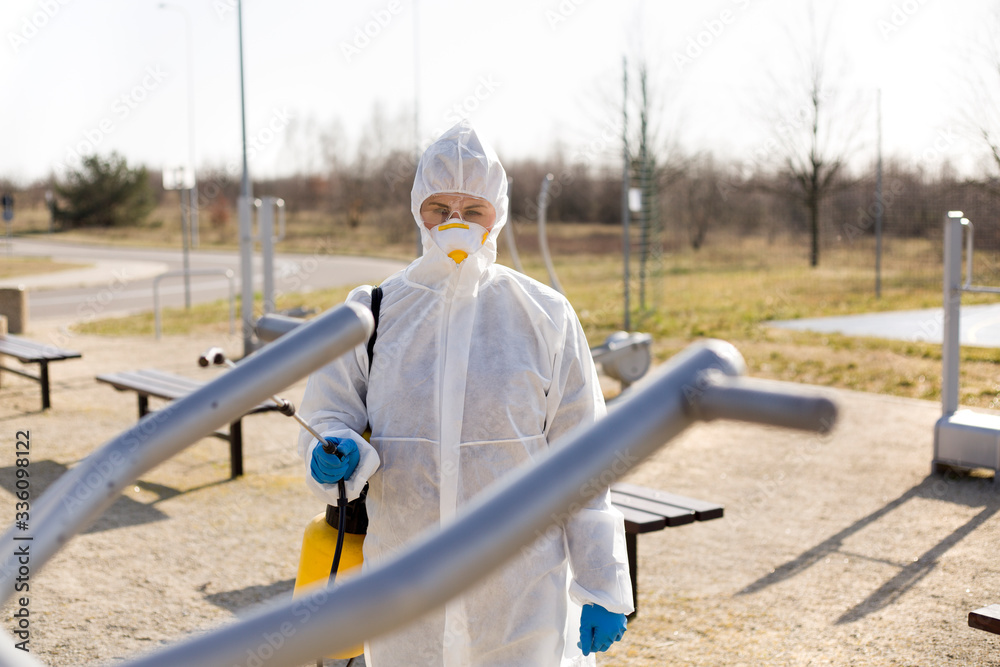 Naklejka premium Osoba w kombinezonie ochronnym spryskuje sprzęt na siłowni zewnętrznej by go zdezynfekować