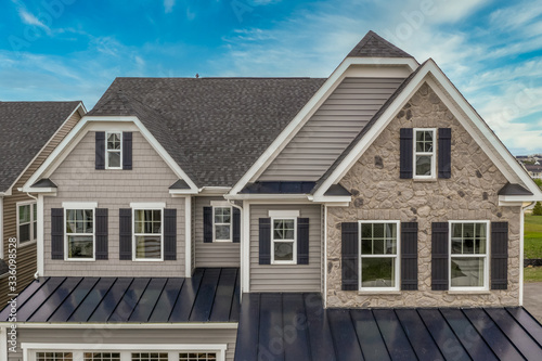 Aerial view of complex facade with stone veneer covered gable 2 jerkin head pitched roof, gray horizontal vinyl siding, dark shutters on the white frame elegant double sash windows single family home